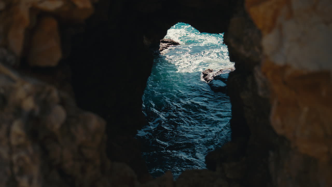 Paredes rochosas costeiras escuras emolduram uma passagem marítima estreita com água azul cintilante, criando uma cena mediterrânea isolada, rica em textura, profundidade e intriga costeira