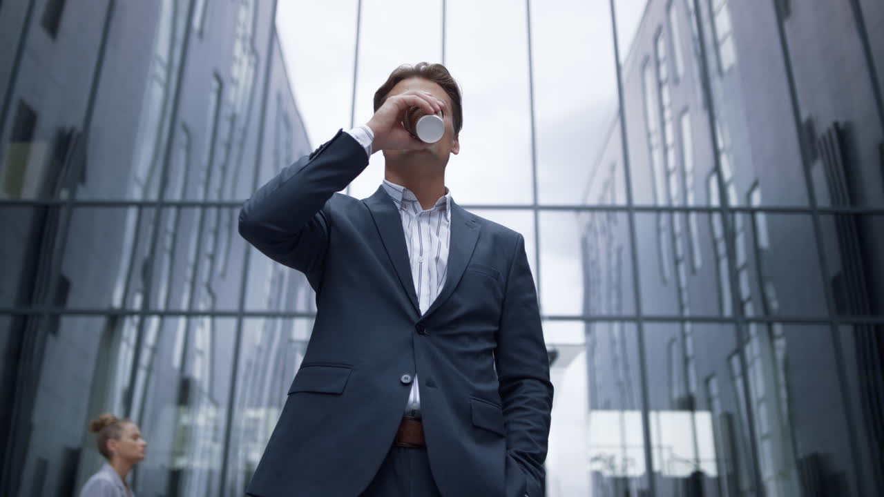 유리 사무실 건물에 서서 커피를 마시는 미소 짓는 사업가의 초상화.