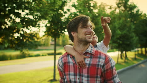 Vater-Und-Sohn-Gehen-Auf-Der-Straße.-Lächelnder-Mann,-Der-Einen-Jungen-Auf-Dem-Rücken-Im-Park-Trägt