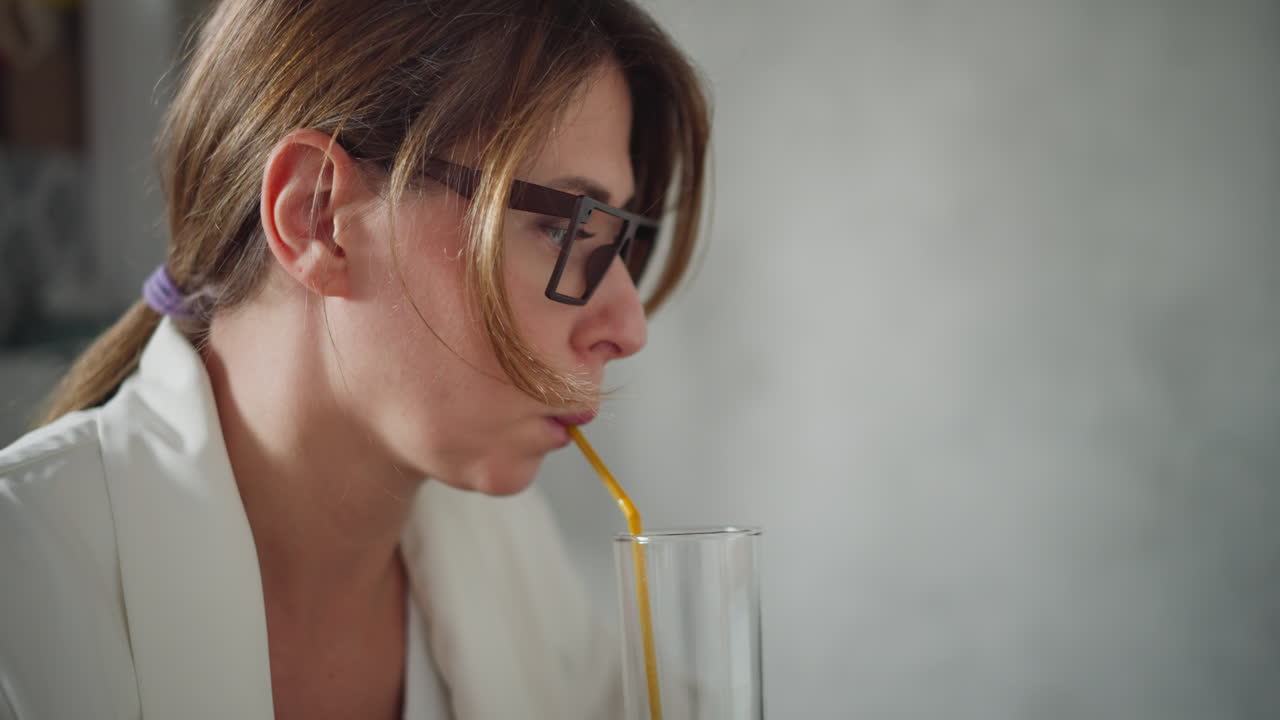 Close up of white lady in white top with hair tied back sipping coffee through yellow straw focused on drink with blur background featuring soft light and unclear item