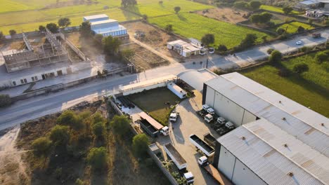 Aerial-view-of-manufacturing-factory-at-sunset-in-india,-sourrounded-by-tropical-landscape-with-plowed-field-and-palm-tree