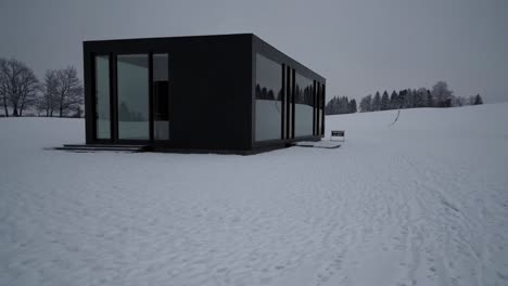 minimalist black tiny house with large windows reflecting snowy landscape, for sale, standing on a snowy field in winter, architecture and real estate concept