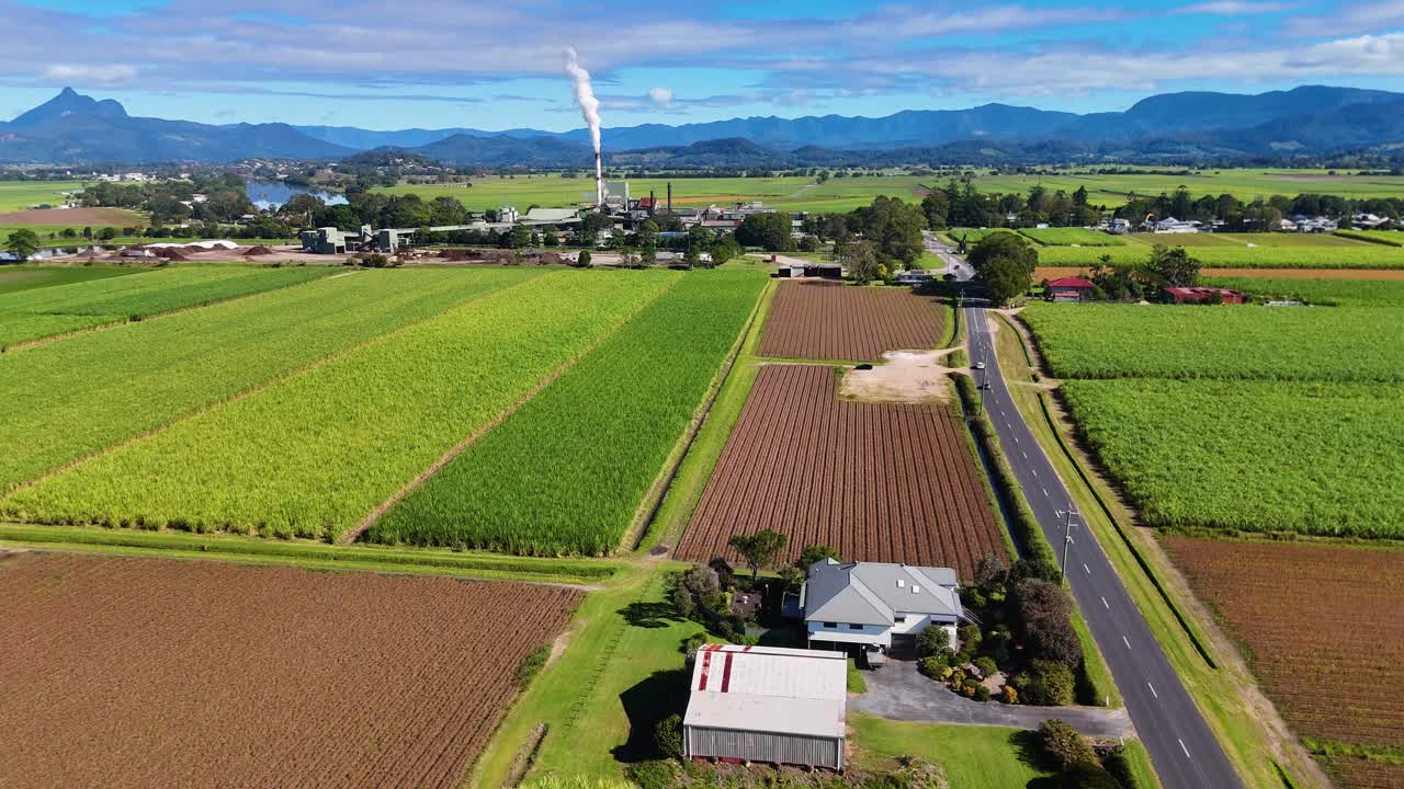 Drone footage captures expansive fields, a road, and a factory emitting smoke under clear skies