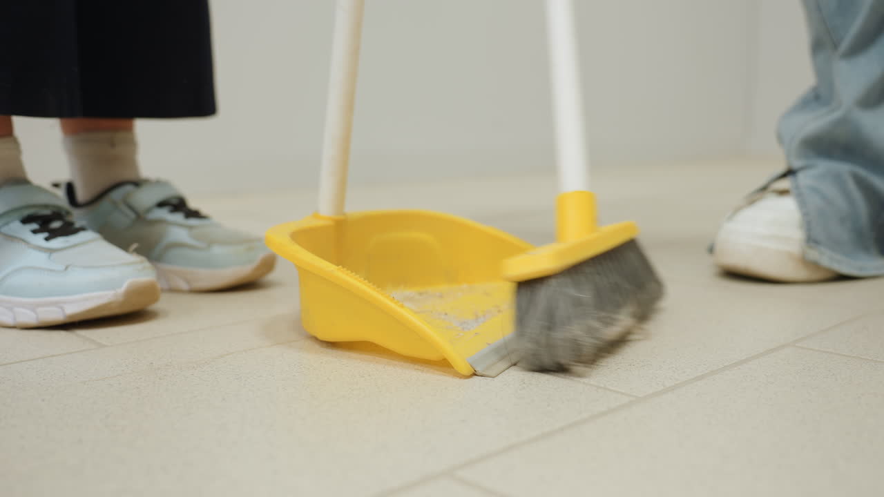 Leg view toddler with parent cleaning tiled floor in laundromat, close sweep action pushing debris into yellow dustpan with broom, teamwork hygiene, sneakers and jeans visible, family chores