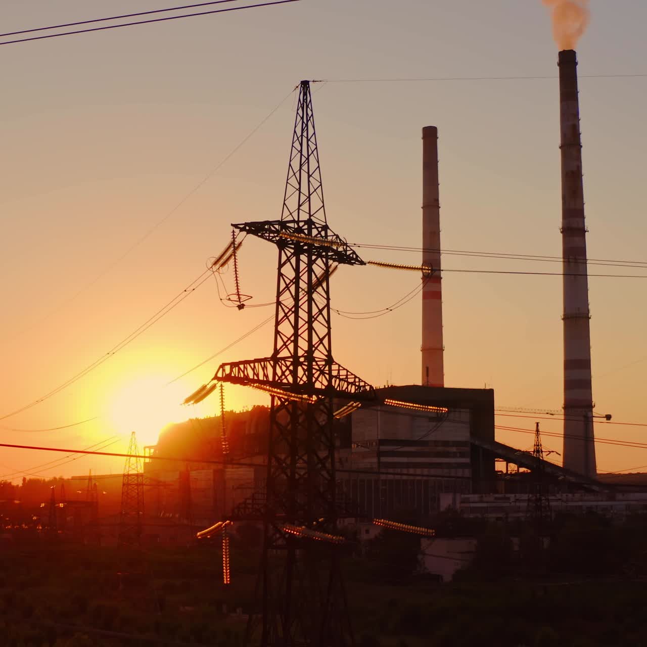 Electricity pylons at sunset. High-voltage electric tower on industrial pipe background. Transmission lines near the factory with dirty smoke into the air in the evening.