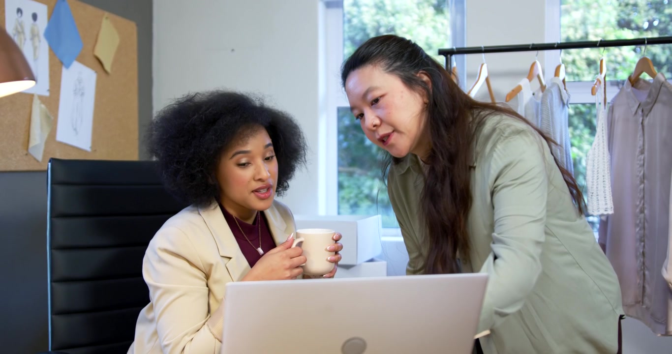 Diverse female team working at desk, standing peer coming after seated peer pointing at laptop, mug