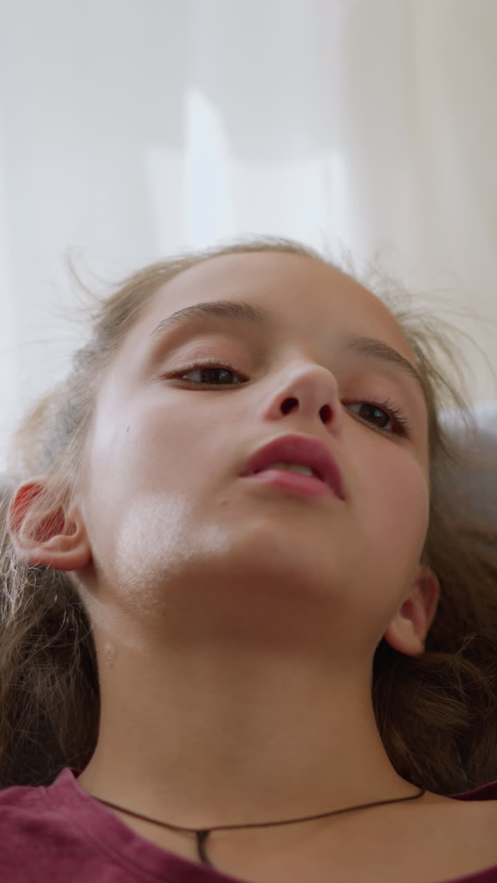 Close-up of little child with red bangle and tied hair sneezing into tissue due to flu, the child appears unwell, lying on the couch with natural light coming through the window