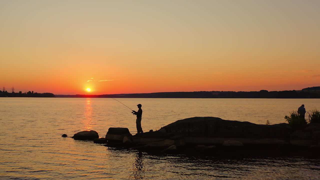 Man fishing at sunset. Silhouette of a man with fishing rod standing on the stone in water in the evening. Fishermen on the lake against orange sunset.