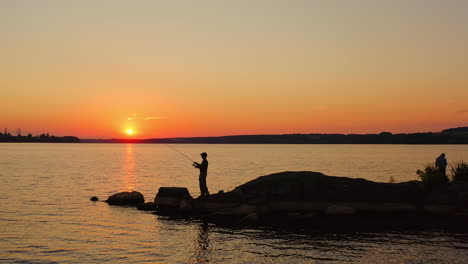 man fishing at sunset. silhouette of a man with fishing rod standing on the stone in water in the evening. fishermen on the lake against orange sunset.