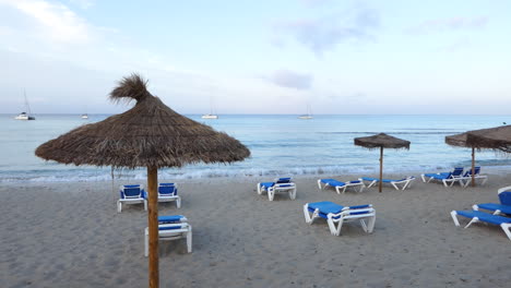 Gran-Vista-De-Una-Playa-Vacía-Al-Amanecer-Con-Tumbonas-Y-Sombrillas-De-Paja,-Pequeñas-Olas-En-El-Mar,-Barcos-Y-Veleros-Anclados