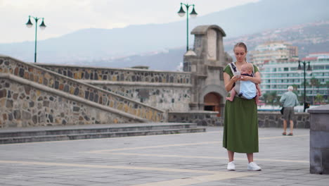 a young mother with her baby in a kangaroo backpack takes photos on a mobile phone while traveling. they walk and glance at the phone screen intermittently