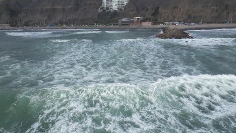 Toma-Aérea-En-Cámara-Lenta-Del-Mar-Con-Olas-Frente-A-La-Playa-De-Miraflores-Lima-En-Perú-Con-Rocas-Y-Edificios-En-El-Fondo