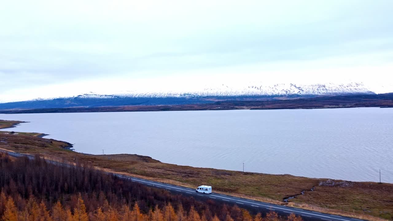 A serene aerial shot captures a motorhome traveling along a road that runs alongside a tranquil lake, reflecting the sky