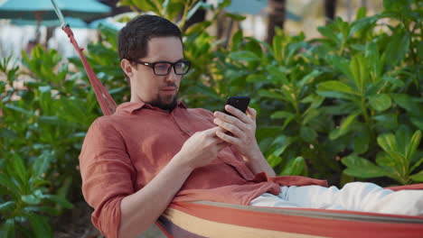hombre relajándose en una hamaca en una playa tropical, usando un teléfono inteligente