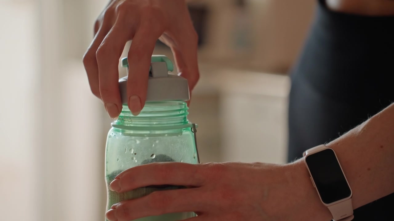 Mujer tapando un vaso mezclador en la cocina después de mezclar. Primer plano de manos con reloj enroscando la tapa en una botella verde, agarre firme y clic audible, luz brillante de la mañana, para fitness e hidratación en casa listos para usar.