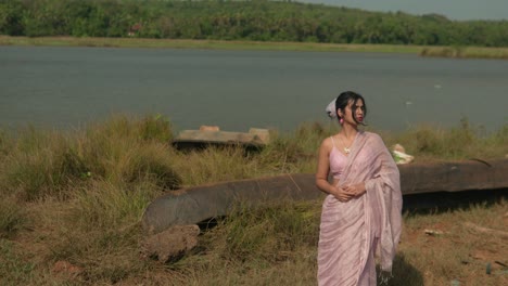 woman in traditional saree by riverside, grass and boat, serene mood, sunny day, static shot