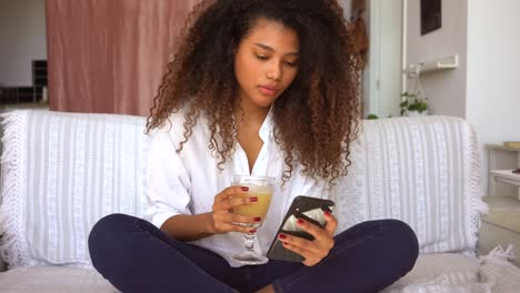 mujer étnica bebiendo jugo y navegando por el teléfono inteligente en casa