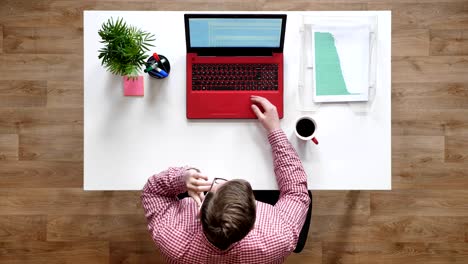 young man in glasses talking on phone, topshot, sitting behind desk with laptop, coffee and documents