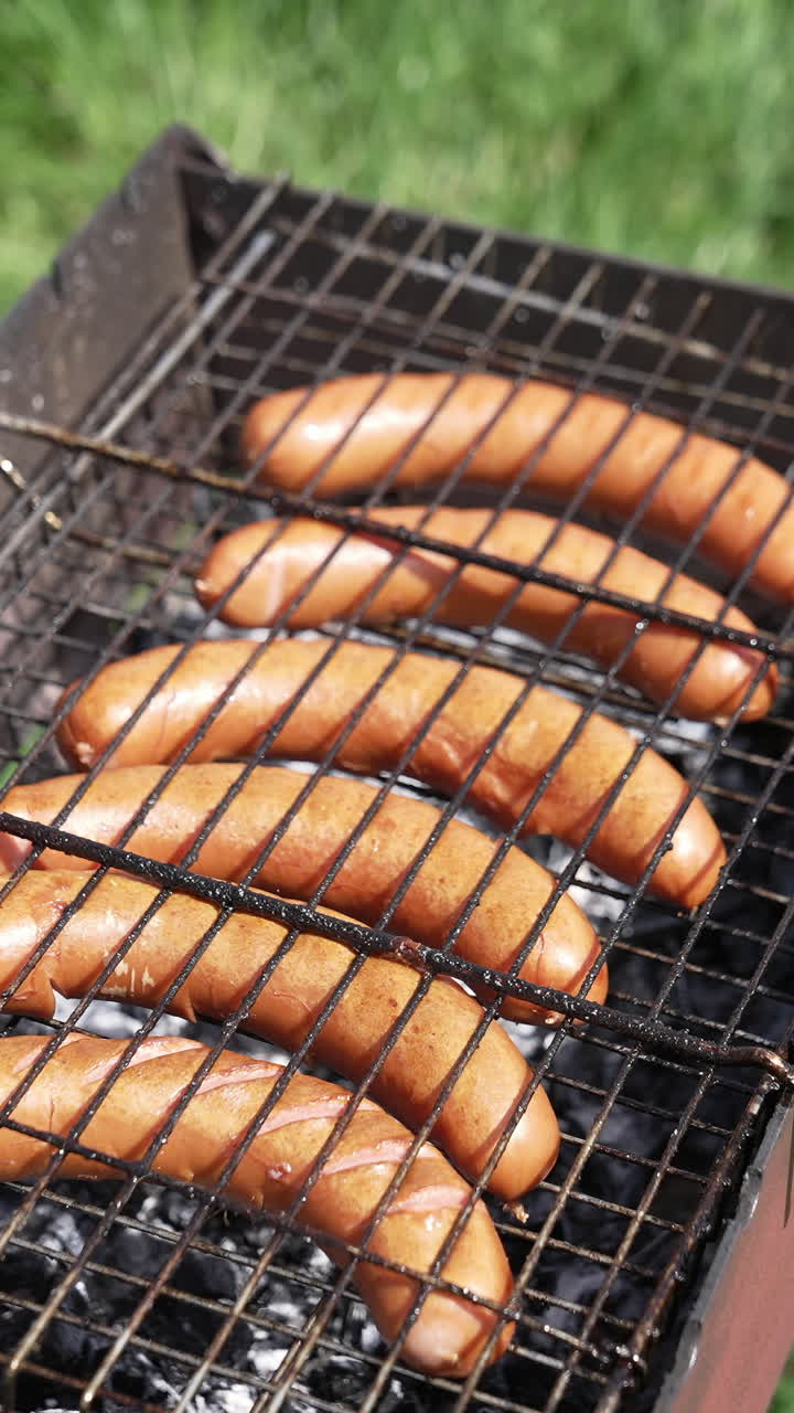Grilling sausage on the grill plate. Grilling food outdoors. Hot sausages roasting in smoke. Barbecue on green grass background. Vertical video