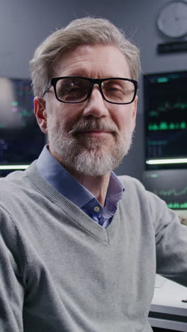 successful mature trader sits near computer with displayed real-time stocks and looks at camera. colleagues analyze exchange market charts at background. concept of cryptocurrency trading. portrait.