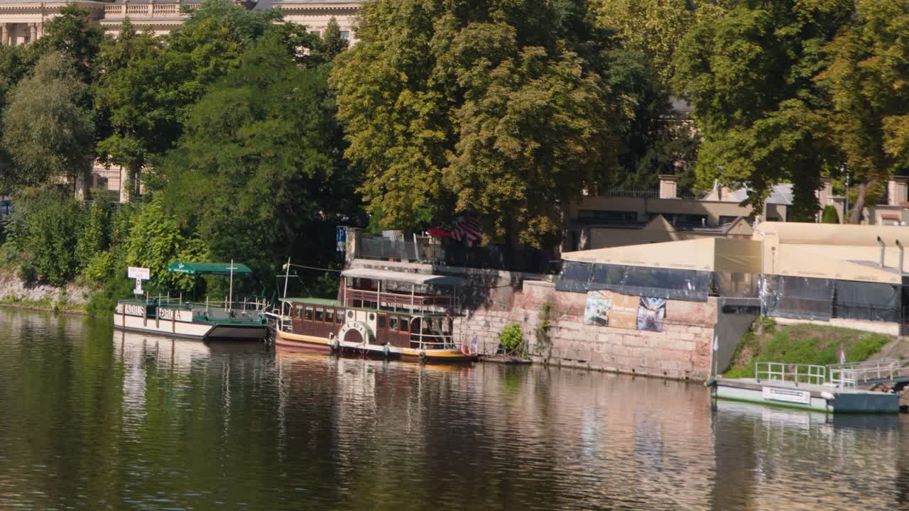 Static wide shot of boats moored along Vltava River, vibrant summer foliage, soft daylight