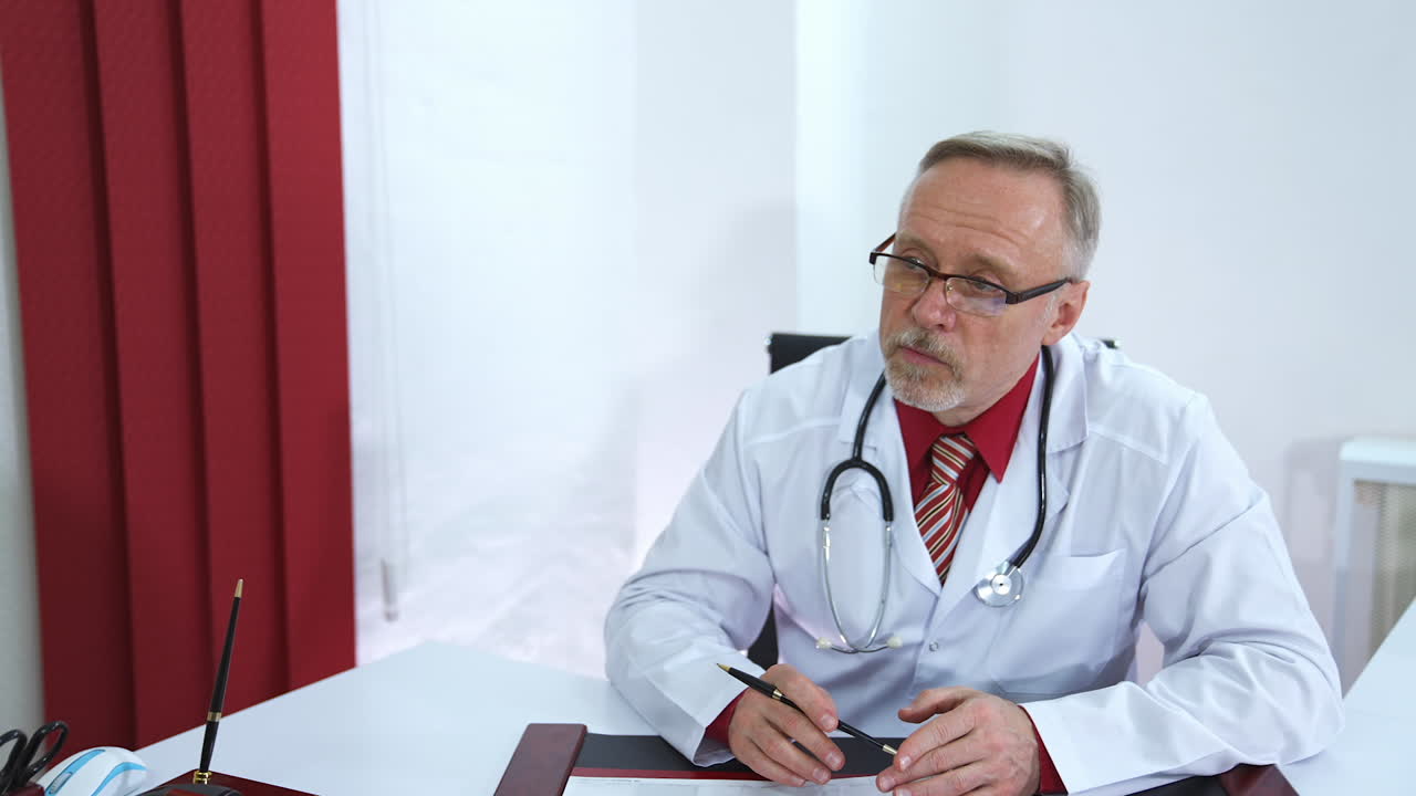 Professional medic sits at table and speaks to a patient. Senior doctor in glasses and grey beard. Phonendoscope on the neck of a phisician.
