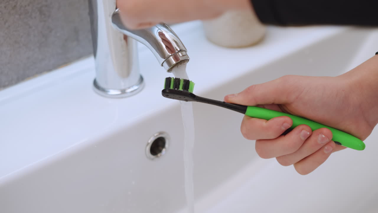 Green toothbrush under chrome faucet rinsing, hand holding toothbrush under running water over porcelain sink, soap dispensers on counter, bristles wet and dripping, morning family hygiene, gentle