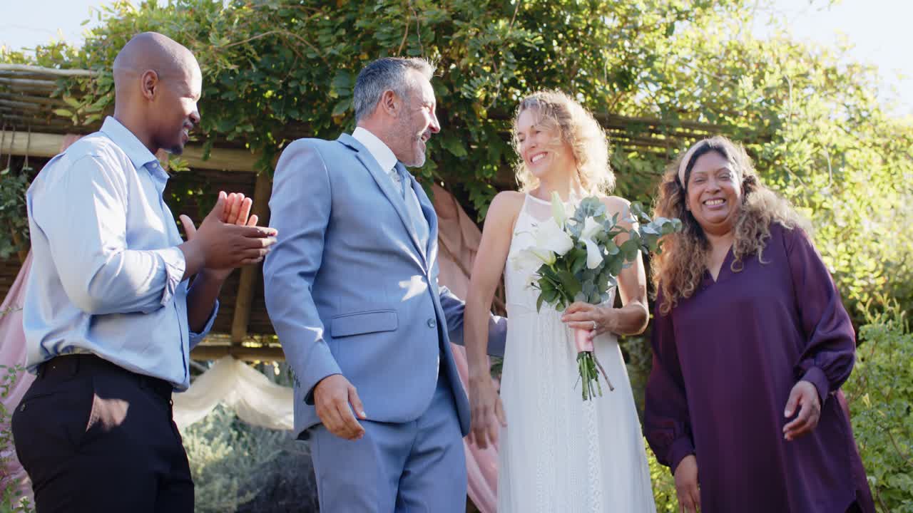 Gruppo eterogeneo sotto una pergola che abbraccia la sposa invitata con il bouquet, il partner si unisce scendendo e festeggiando.