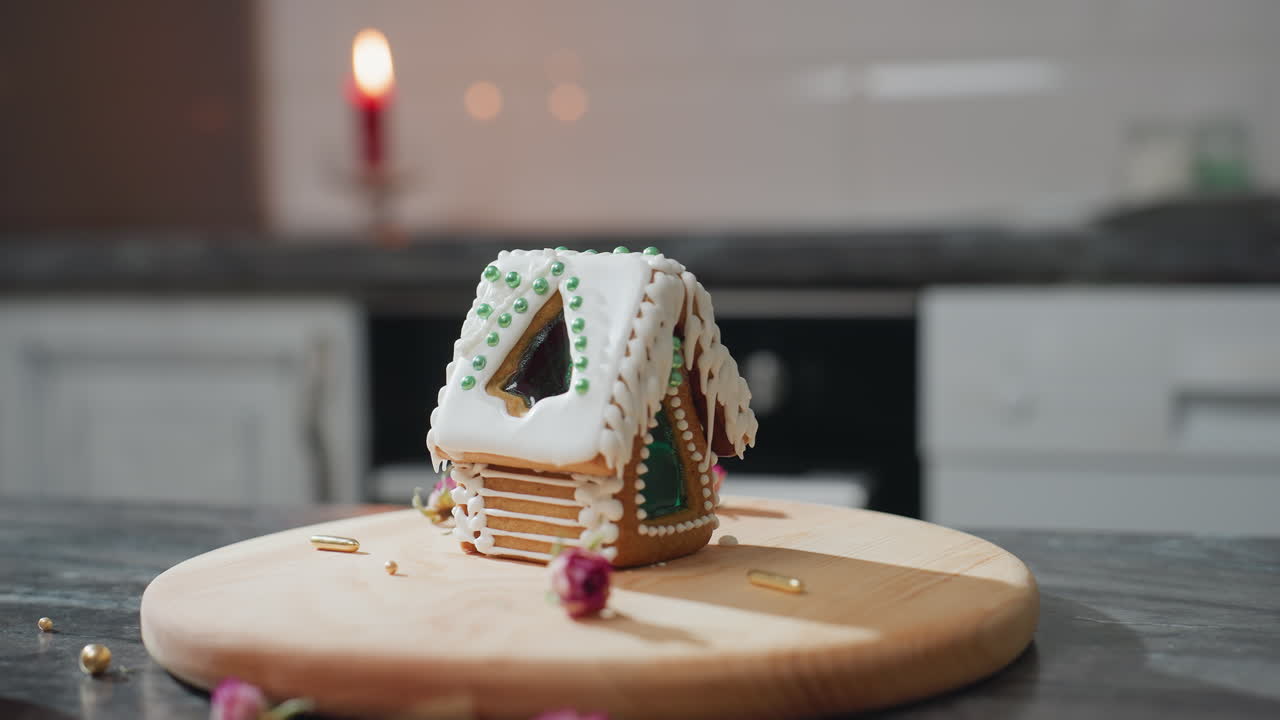 una casa de pan de jengibre bellamente decorada con hielo blanco y cuentas verdes se sienta en una bandeja de madera en una cocina acogedora, a la luz de velas cálidas, cuentas doradas esparcidas y flores secas