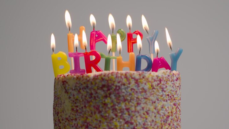 Lit Candles Spelling Happy Birthday On Cake Being Blown Out Against Grey Studio Background