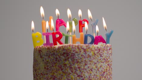 Lit-Candles-Spelling-Happy-Birthday-On-Cake-Being-Blown-Out-Against-Grey-Studio-Background