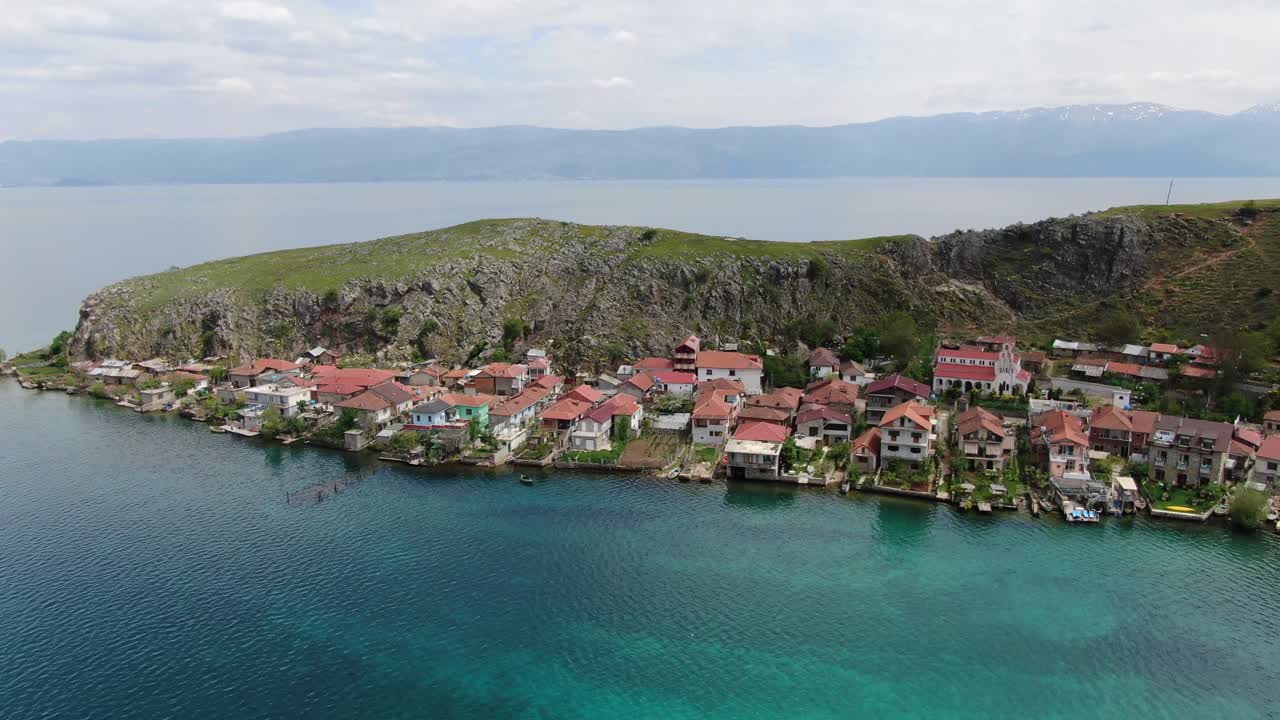 Arnavutluk'ta, berrak mavi suya sahip Ohri Gölü kenarında yeşil bir tepenin bitişiğindeki evlerin bulunduğu küçük bir kasabanın üzerinden uçan drone görüntüsü.