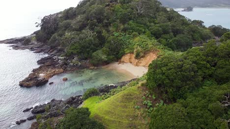 Rising-aerial-drone-of-green-rocky-outcrop-in-the-rain-on-Maitai-Bay-in-the-rain,-Karikari-Peninsula,-Northland,-New-Zealand-Aotearoa