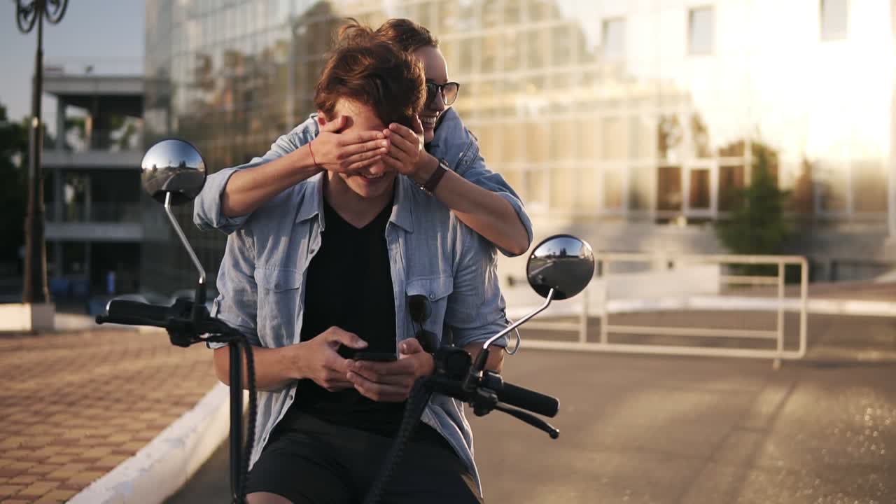 Young caucasian man is sitting on bike and scrolling his mobile phone. The attractive girlfriend coming from the back and closed his eyes with hands. Guess who. Happy, cheerful young people outdoors