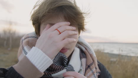 close up view of teenage girl with short hair and winter clothes looking at camera and smiling on seashore on windy day