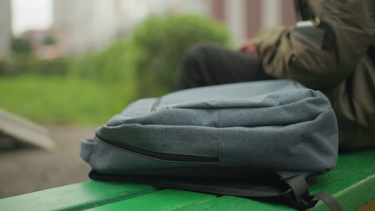 Close up of school bag placed on green bench outdoors with kid seated beside holding handheld device, blurred grassy background creating calm and casual everyday atmosphere
