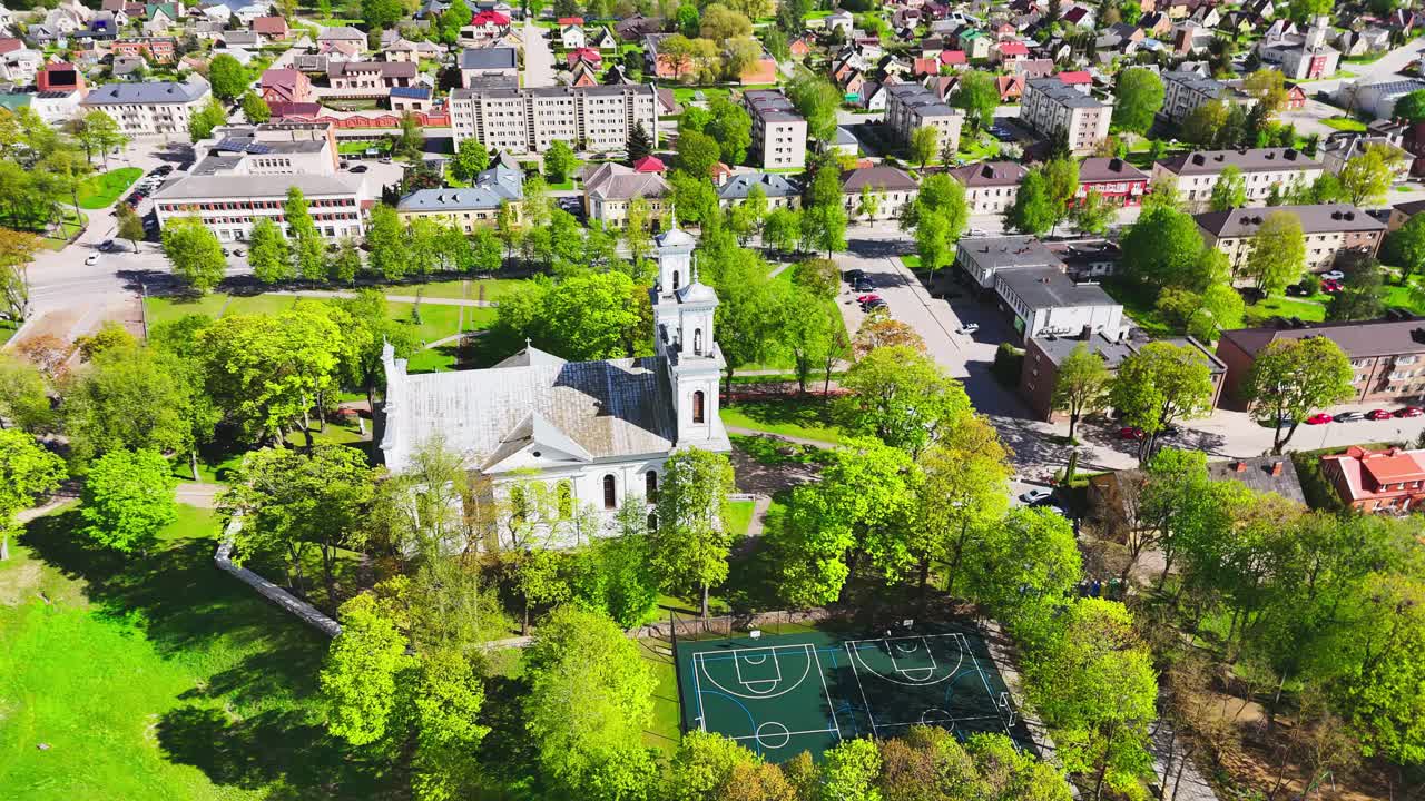 An old church with twin towers stands at the heart of a residential neighborhood, encircled by lush trees and a nearby basketball court under bright daylight.