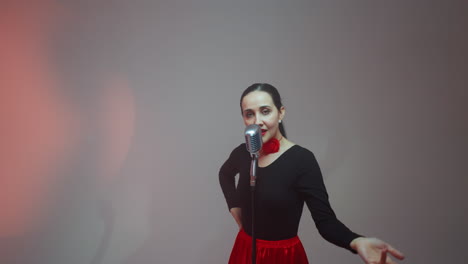 vocalist in a black and red gown standing in front of a vintage mic moving hands slowly, guitarist playing a red guitar, and keyboardist playing a red sampler against a grey background