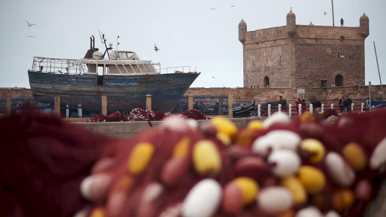 Camera slides right with out of focus fish nets in foreground and fishing boats in focus background