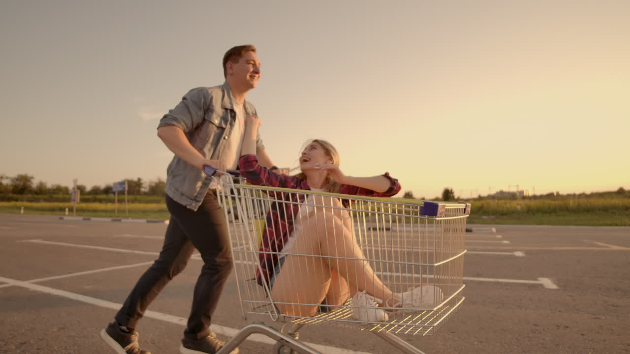 joven coupé elegante divirtiéndose montando con un carrito de compras en el estacionamiento al aire libre cerca del supermercado