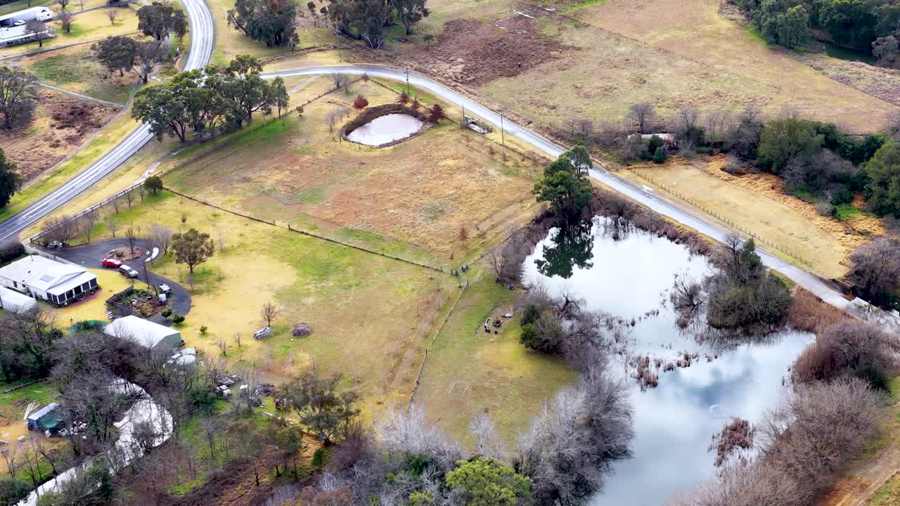 Drone footage smoothly glides above open farmland with ponds, scattered trees, rural homes, and winding roads under soft daylight, capturing a peaceful countryside landscape