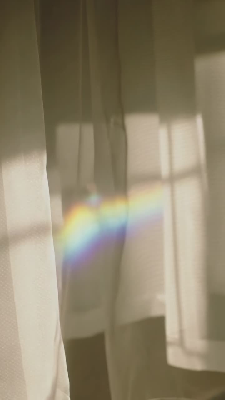 Vídeo vertical: Sol baixo refratando faixa de arco-íris pela cortina na janela, mostrando sombras dos montantes