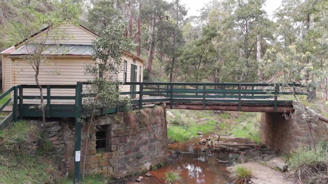 el dron vuela hacia un puente de madera y piedra sobre un pequeño arroyo, con un cobertizo de tablones de madera a la izquierda