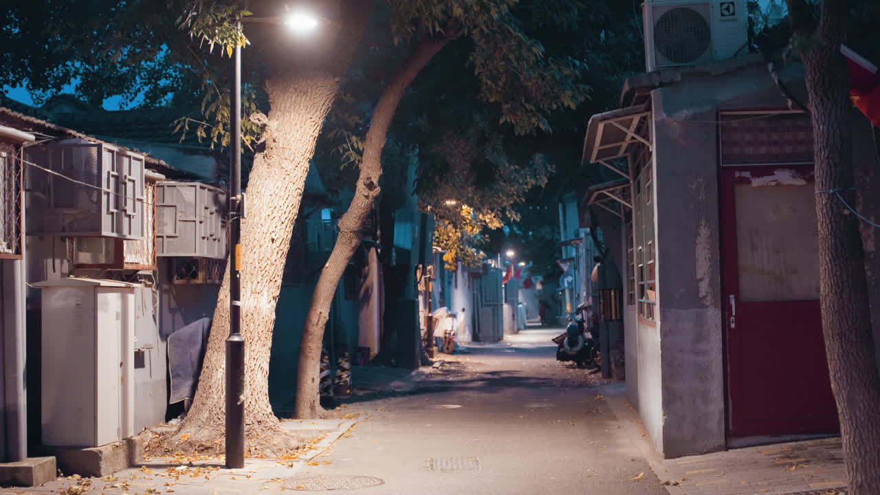 Night alley empty under warm lamp, leafstrewn pavement flanked by old shopfronts and bowed trees, soft amber glow mixing with cool teal background, cinematic stillness and nostalgic atmosphere, slow