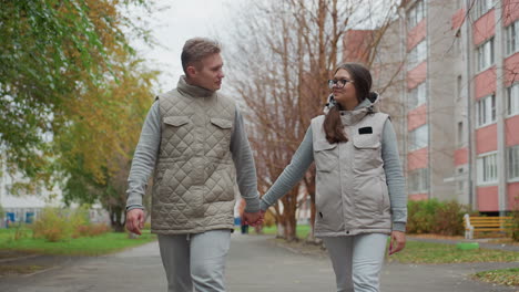 joyful moment of young couple walking hand in hand, swinging arms while smiling and looking at each other with affection during peaceful stroll along tree-lined pathway with blurred urban background