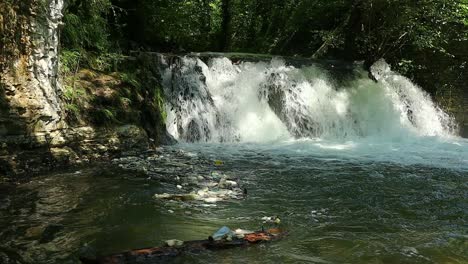 garbage-floating-at-the-foot-of-a-beautiful-waterfall-in-a-relaxing-forest-landscape,-slowmotion-shot