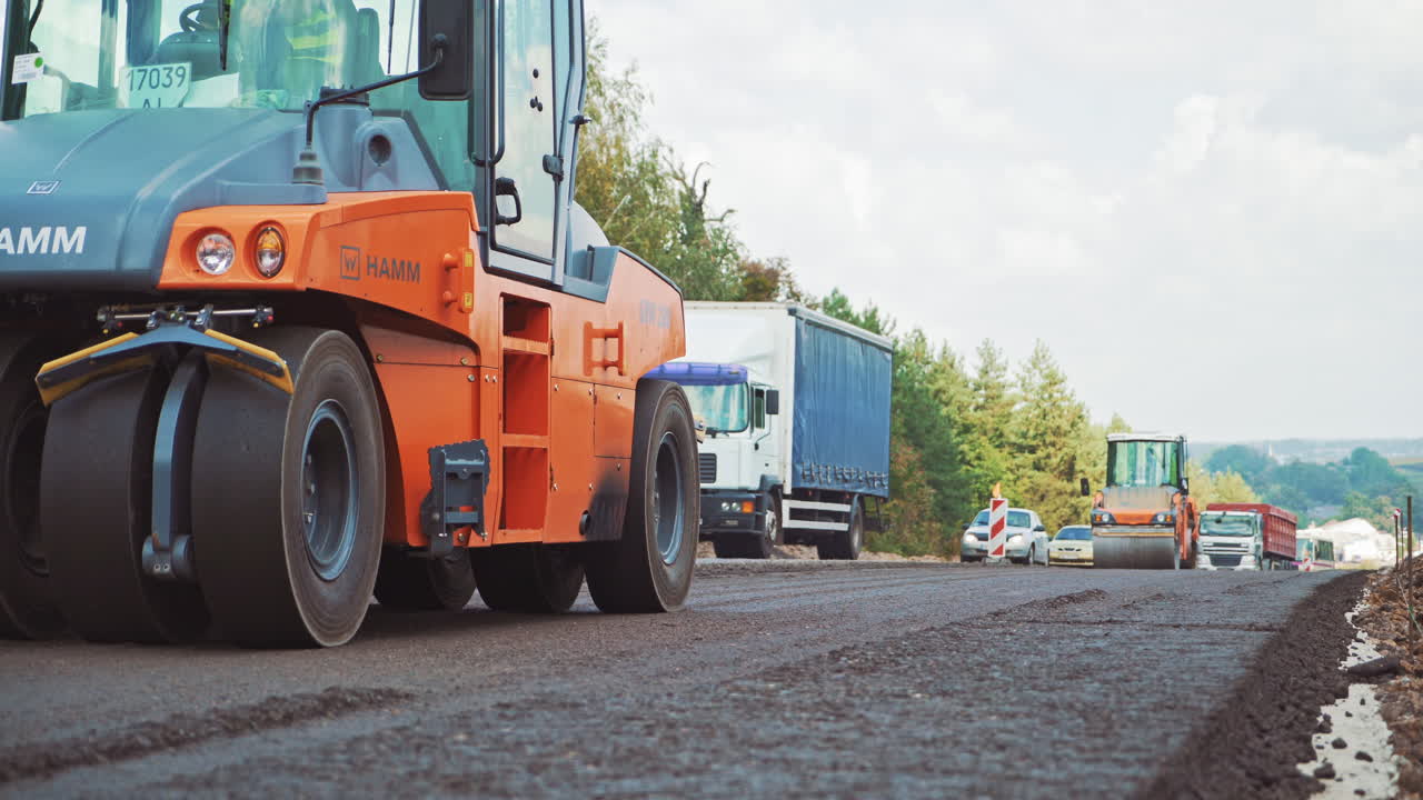 Orange roller machine pressing hot asphalt on the road. Two compactor machines are making new asphalt on the background of traffic movement. Street resurfacing.