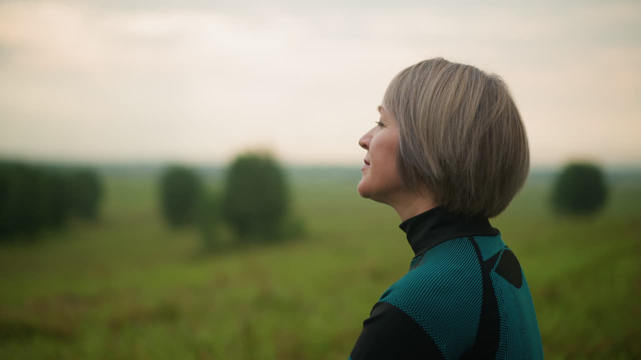 mujer de traje verde y negro mirando a la distancia con una sonrisa suave, en un campo de hierba brumosa, árboles borrosos en el fondo