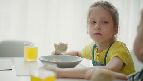close up of young girl wearing yellow top sitting at dining table eating lunch with large dumpling on fork, focused expression, glass of fruit juice beside her on bright naturally lit table