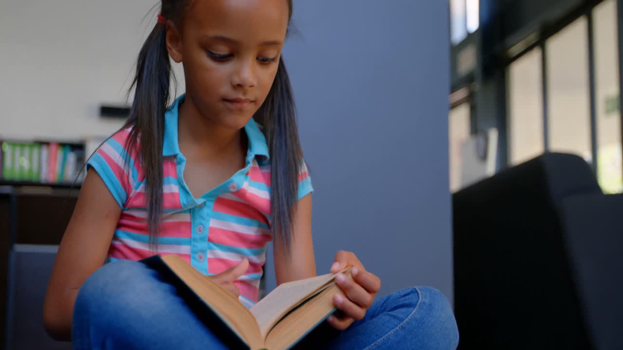 Front view of attentive African American schoolgirl reading a book in library at school 4k